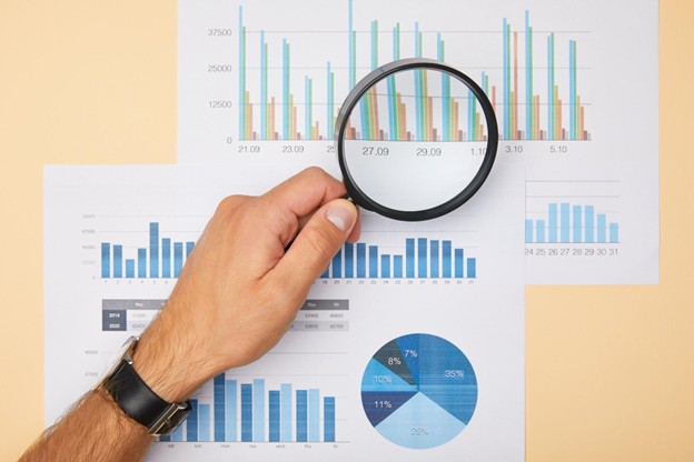 A hand holding a magnifying glass up to several papers containing an array of charts and graphs.