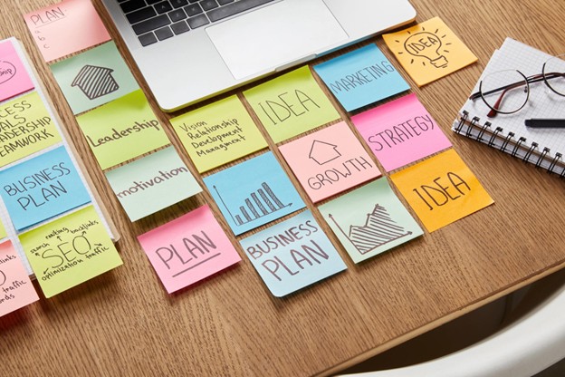 An arrangement of colorful sticky notes on a table, each including key phrases related to business plans, such as SEO, growth, and marketing. A laptop and pair of glasses are also on the table.